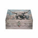 A rectangular wooden storage box with a rustic weathered wood grain finish, featuring a printed illustration of a shaggy Highland cow in a field of pink flowers on the lid. The front has a decorative silver-toned metal clasp with a red tassel, set against a plain white background.