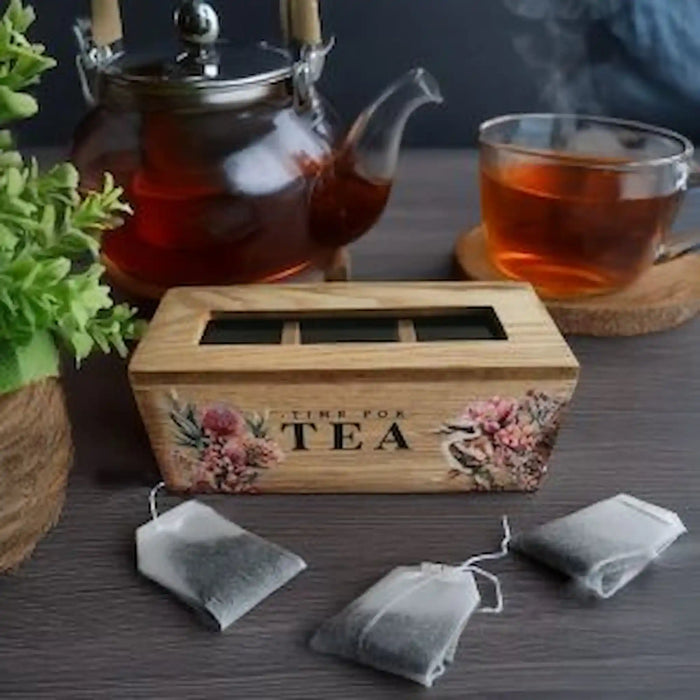 A wooden tea box with floral illustrations and the words 'TIME FOR TEA' sits on a dark tabletop, with tea bags in front and a glass teacup and steaming teapot in the background.