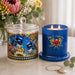 A decorative blue scented candle in a glass jar with a matching lid, shown beside the unlit candle on a light marble surface. The lid features a colorful illustration of blue birds and floral motifs, while the candle jar is deep cobalt blue with gold script reading 'Joy & Happiness' and a floral design.