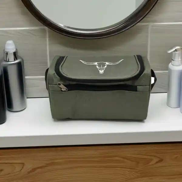 An olive green toiletry bag with a silver bull skull logo, resting on a white bathroom counter with toiletry bottles and a mirror in the background.