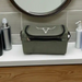 An olive green toiletry bag with a silver bull skull logo, resting on a white bathroom counter with toiletry bottles and a mirror in the background.