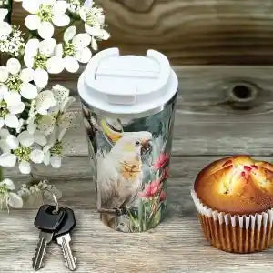 A travel tumbler with a white lid and a printed bird-and-floral design, shown on a rustic wooden surface beside white flowers, keys, and a muffin.