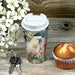 A travel tumbler with a white lid and a printed bird-and-floral design, shown on a rustic wooden surface beside white flowers, keys, and a muffin.