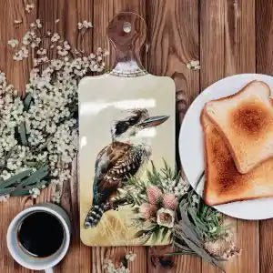 A rectangular wooden cutting board with a handle, featuring a printed illustration of a kookaburra bird and small flowers, styled on a wooden table with a slice of toast on a plate, a cup of coffee, and dried flowers.