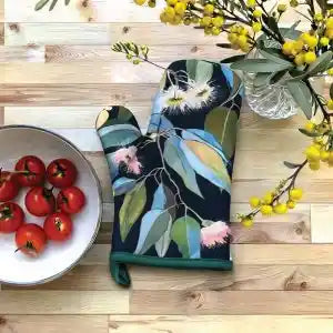 A floral oven mitt with a navy blue background, green leaves, and pink flowers, styled on a light wood surface next to a bowl of red tomatoes and yellow flowers.