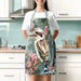 A woman wearing a white t-shirt and a floral kookaboo print kitchen apron, standing in a kitchen setting.