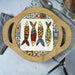 A round wooden serving tray with two side handles, featuring a colorful Mediterranean-style fish pattern in the center, set on a light gray tabletop with kitchen utensils.