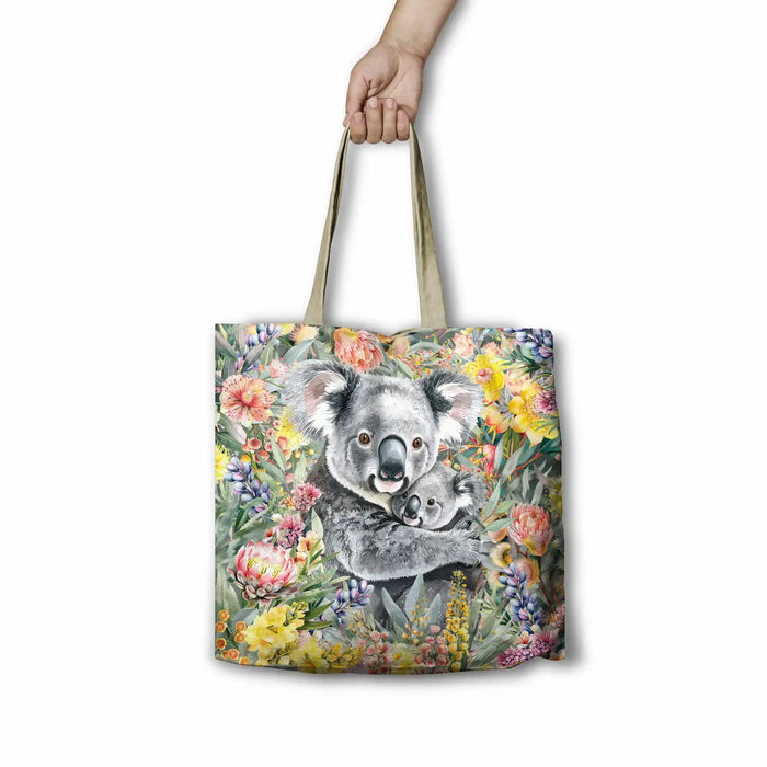 A tote bag with a printed illustration of a koala and its joey surrounded by colorful flowers, held by a hand against a plain white background.