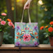 A tote bag with a colorful floral and bird print is held by a hand and placed on a wooden surface outdoors, flanked by potted flowers.