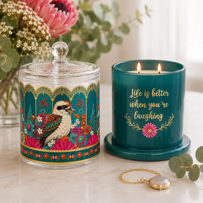 A decorative teal candle jar with a glass lid and matching base sits beside a teal candle in a lit holder. The lid features a colorful kookaburra illustration with floral and geometric border details.