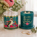 A decorative teal candle jar with a glass lid and matching base sits beside a teal candle in a lit holder. The lid features a colorful kookaburra illustration with floral and geometric border details.