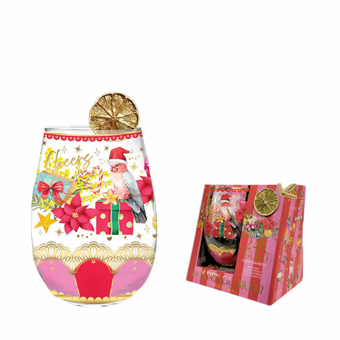 A clear stemless wine glass with a festive Christmas design, shown next to its pink gift box. The glass features a colorful illustration of a bird, a gift box, and the words 'Merry Christmas' decorated with gold stars and red flowers.