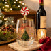 "Merry Grinchmas!" stemless wine glass with peppermint swirl bottle stopper on a marble coaster with a wine bottle, candle, pine cones and red baubles