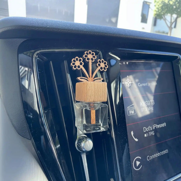 A wooden flower-shaped car air freshener with three flowers and a clear glass bottle filled with amber liquid, clipped onto a car dashboard air vent next to the infotainment screen.