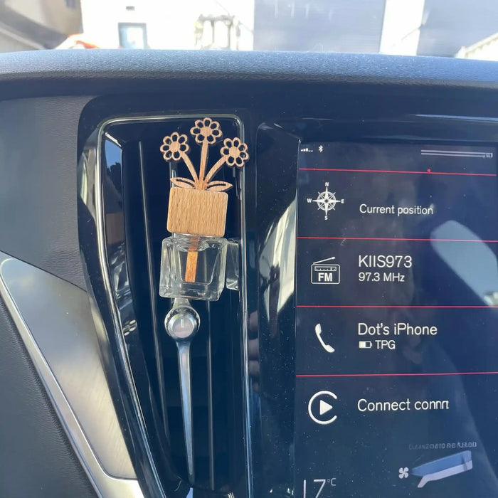 A car air freshener with a wooden flower bouquet and clear bottle, inserted into the air vent of a vehicle dashboard next to the infotainment screen.