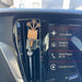 A car air freshener with a wooden flower bouquet and clear bottle, inserted into the air vent of a vehicle dashboard next to the infotainment screen.