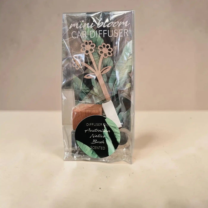 A packaged mini bloom car diffuser with a clear plastic bag, showing the wooden flower-shaped diffuser, sponge, and scent card on a light beige surface against a neutral beige background.