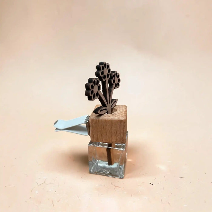 A small decorative hair clip or hair accessory with a clear glass square base, a light blue metal spring, and a dark brown wooden top featuring a flower-shaped design, set against a solid beige background.