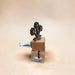 A small decorative hair clip or hair accessory with a clear glass square base, a light blue metal spring, and a dark brown wooden top featuring a flower-shaped design, set against a solid beige background.