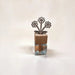 A small decorative object with a clear glass base, a light wood cap, and a metal flower-shaped top, photographed on a plain light background.