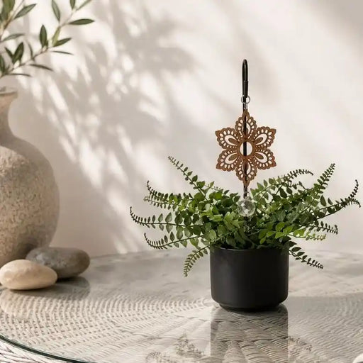 A decorative laser-cut metal winter ornament in a warm brown tone hangs above a small potted fern on a glass table, with a ceramic vase and stones in the softly lit background.
