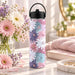 Quiet Blossom 620ml insulated water bottle with pastel daisy artwork, styled beside fresh flowers and a pink towel.