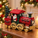 A decorative Christmas steam train figurine sits on a wooden table in a warmly lit holiday setting. The train is primarily red and dark green with gold-toned wheels and trim, featuring Santa Claus in the driver's seat, a small Christmas tree, a teddy bear, and gift boxes.