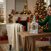 A woman in a green sweater places a small white Christmas church ornament on a wooden table in a cozy, warmly lit living room decorated for Christmas.