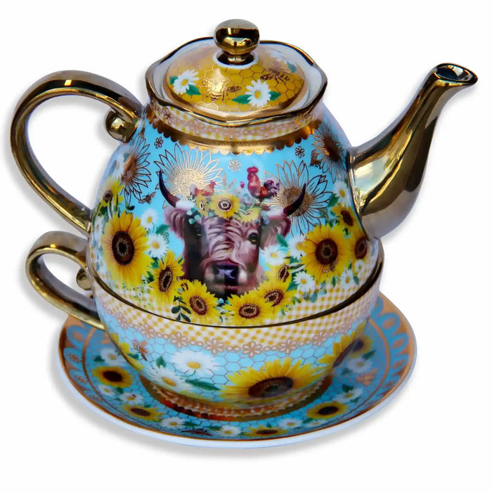 An ornate ceramic tea-for-one set featuring a teapot stacked on a matching cup and saucer, decorated with a highland cow, sunflowers, daisies, bees, and gold honeycomb detail on a sky blue background with gold trim