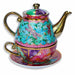 An ornate ceramic tea-for-one set with a teapot stacked on a matching cup and saucer, decorated with purple and pink dragonflies, teal hydrangea florals, and patchwork panels in pink, purple, and teal with gold honeycomb trim