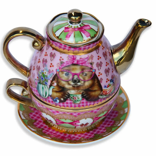 A pink and gold porcelain tea for one teapot with a matching cup and saucer, decorated with a whimsical Wonder Wombat illustration wearing pink glasses and a bow, on a white background.