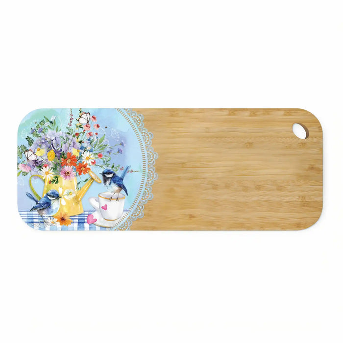 A rectangular wooden cutting board with rounded corners and a small oval hole in the top right. The left side features a decorative printed illustration with a bouquet of flowers, a yellow teapot, a teacup, bluebirds, and a lace border over a light blue background.