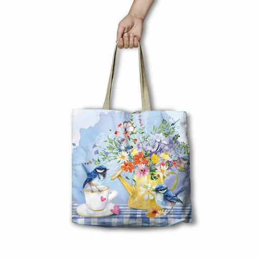 A tote bag with a watercolor-style floral and bird print, held by a hand against a plain white background. The print features blue birds, a yellow teapot filled with multicolored flowers, a teacup, and a blue-and-white checkered surface.