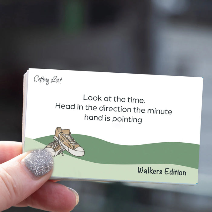 A hand with glittery nail polish holds a square card that reads 'Look at the time. Head in the direction the minute hand is pointing' with 'Getting Lost' at the top and 'Walkers Edition' at the bottom.