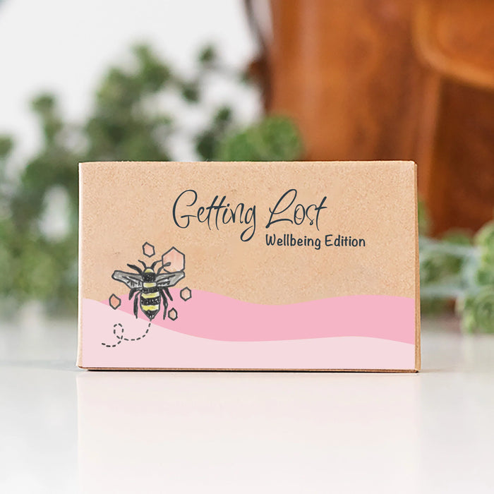 Kraft paper box with a bee illustration and 'Getting Lost Wellbeing Edition' text on a blurred background.