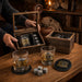 A whiskey gift set displayed on a wooden table in a cozy room setting. The set includes two clear engraved whiskey glasses with black and gold text reading 'CRUMPY'S THE MAN'S CLUB' and 'THE MAN THE MYTH THE LEGEND', two dark gray stone cubes, a round black coaster, and two wooden presentation boxes containing additional glasses.
