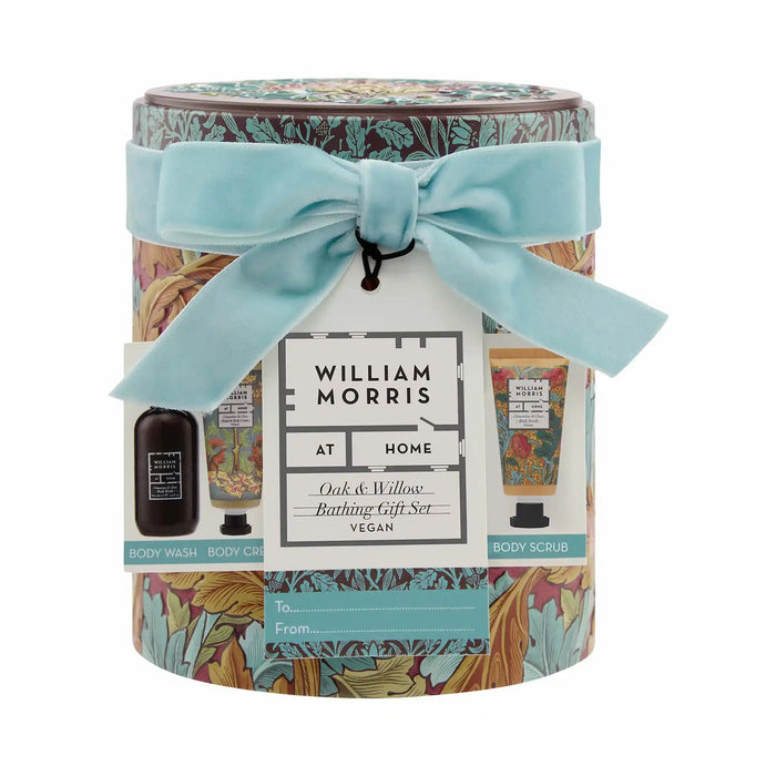 William Morris At Home Oak & Willow Bathing Gift Set in a cylindrical tin with a light blue velvet ribbon bow. The tin features a detailed botanical pattern and a white hanging tag showing product images of a body wash, body cream, and body scrub.