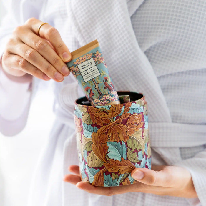 A person in a white robe holds a decorative cylindrical container with an ornate botanical pattern while inserting a matching patterned tube into it.