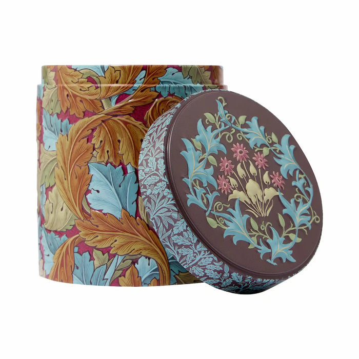 A round metal tin with a floral and botanical pattern, shown with its lid leaning against it. The tin features a multicolor design with blue, gold, and burgundy leaves, and the lid has a dark brown background with a matching floral motif in blue, pink, and gold.