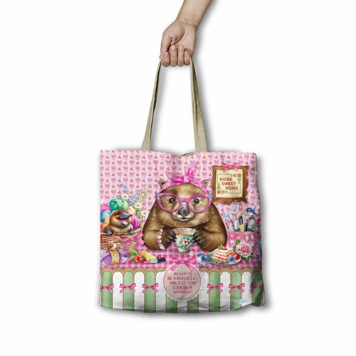 A tote bag with a whimsical illustration of a brown bear wearing pink glasses and a bow, holding a teacup on a pink patterned background. A hand is holding the tan shoulder straps of the bag against a plain white background.